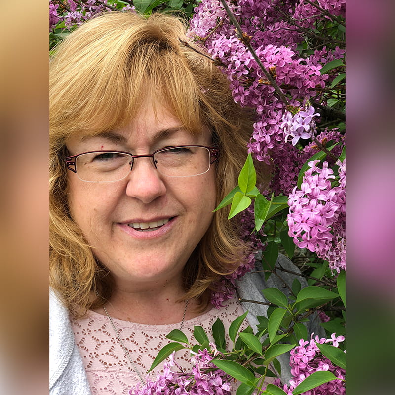 white middle aged woman with auburn hair and glasses smiling next to lilacs in bloom