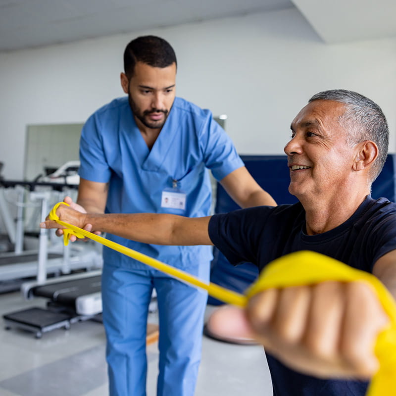 Mature Latin American man doing physical therapy exercises using a stretch band with the assistance of his therapist