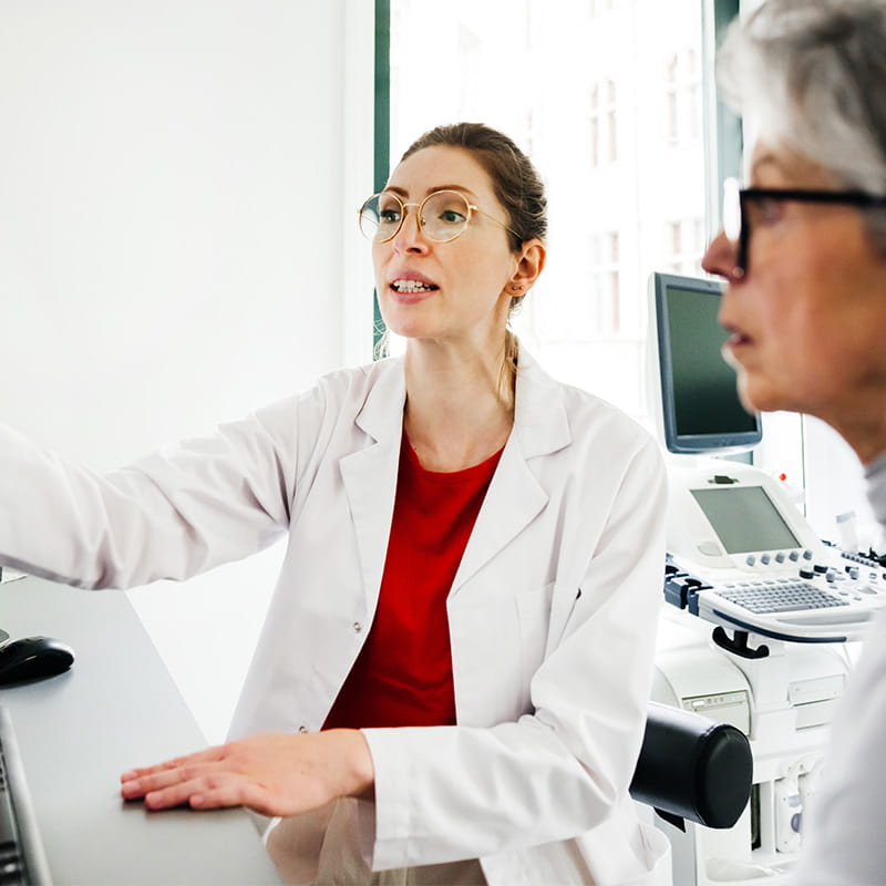 A young female doctor points to a computer monitor with CT scans while explaining test results to a senior female patient seated beside her.