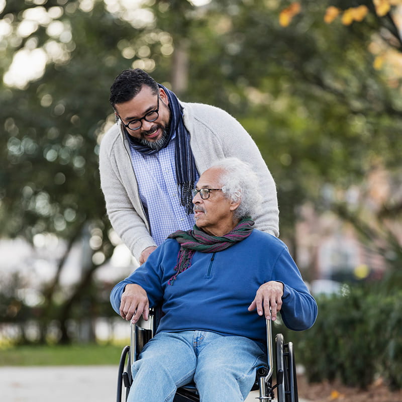 A senior Hispanic man in his 80s sitting in a wheelchair, taking a walk in the park with his adult son, a mid adult man in his 30s.