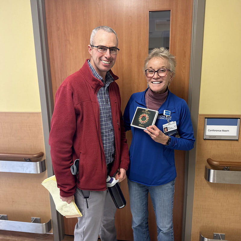 A white man wearing glasses and dressed in a maroon jacket stands next to a white female doctor wearing a blue sweater. Both are smiling. The doctor is holding a Christmas music CD.