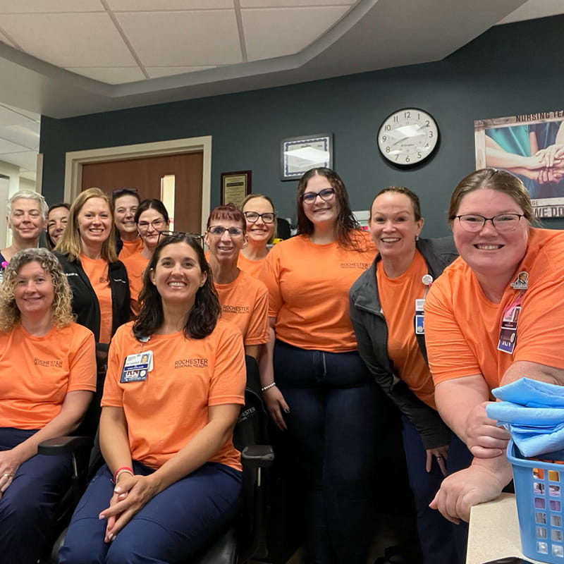 Three rows of healthcare workers in orange shirts smile in an office