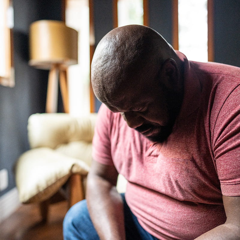 Sad middle aged Black in maroon t-shirt looking down while sitting in living room