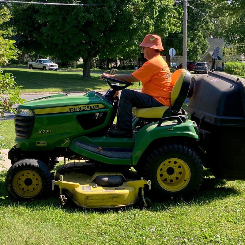 Man in orange shirt and orange hat driving green and yellow lawnmower on green grass