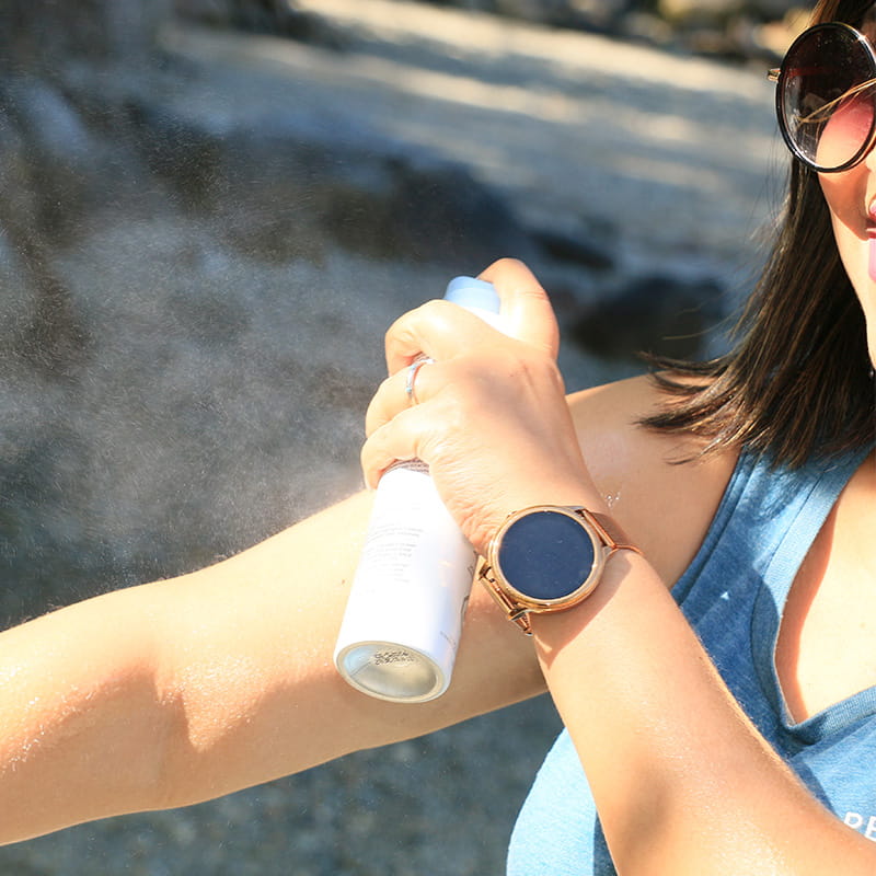 A woman spraying sunscreen on her bare arm. She is wearing sunglasses, a blue sleeveless top, a watch, bracelet, and holding an aerosol can of sunscreen
